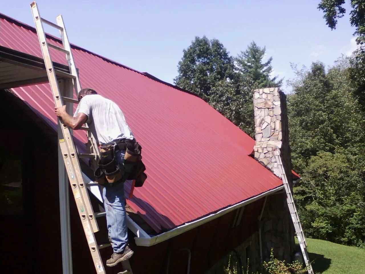Roofer on a ladder beside a red metal roof and stone chimney — Soffit & Fascia Repair by Roofing Brothers in Cairo